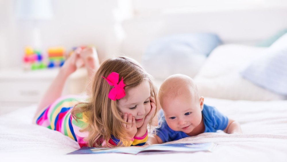 funny happy toddler girl reading a book and playing with newborn baby boy in bed. kids play at home. white nursery. child in sunny bedroom. children read and study. interior for baby and young kid.