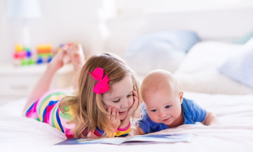 49114765 - funny happy toddler girl reading a book and playing with newborn baby boy in bed. kids play at home. white nursery. child in sunny bedroom. children read and study. interior for baby and young kid.